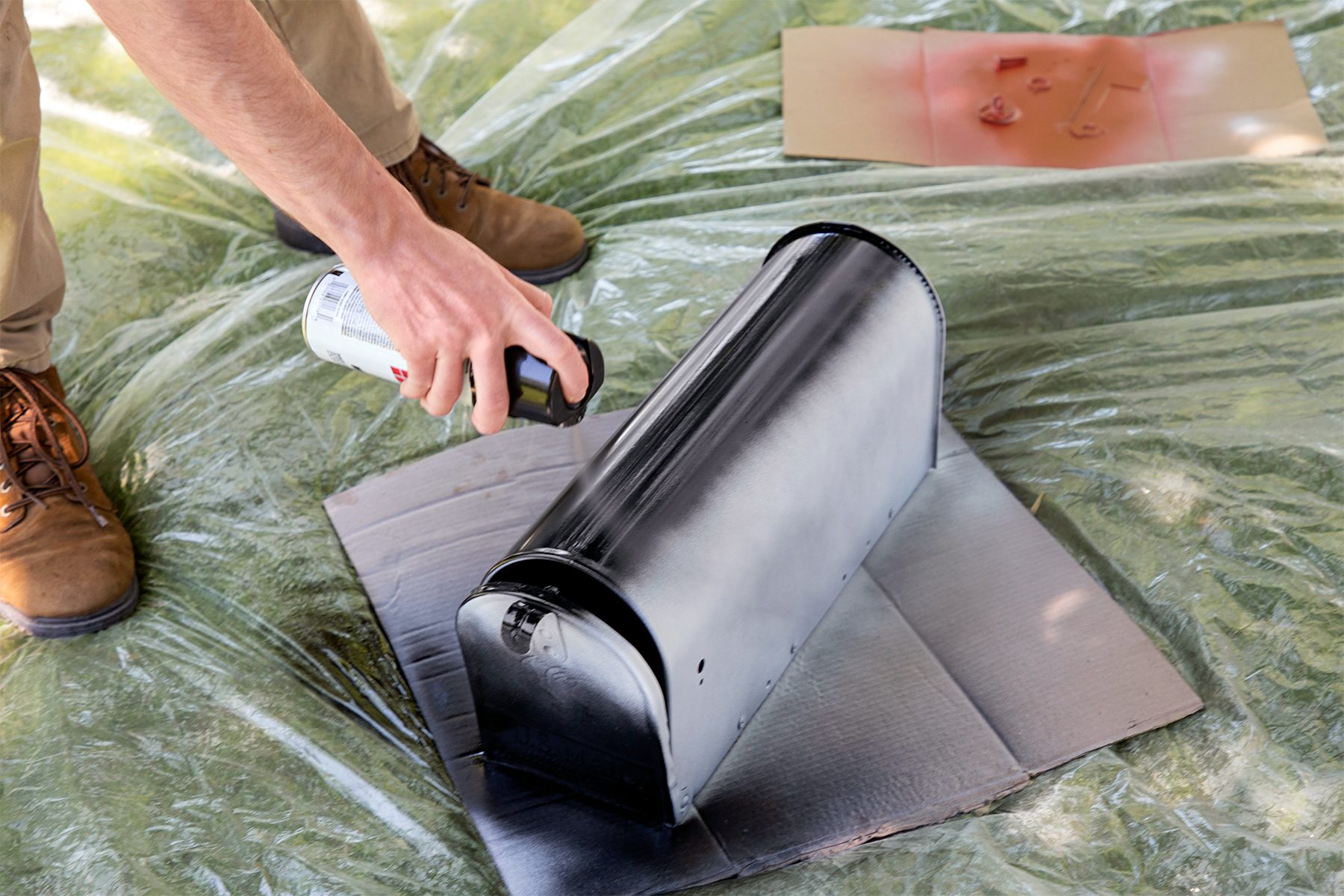 A person wearing brown shoes and beige pants is spray painting a black mailbox on cardboard sheets, with clear plastic covering the surrounding area. In the background, there is a paint-splattered piece of cardboard.