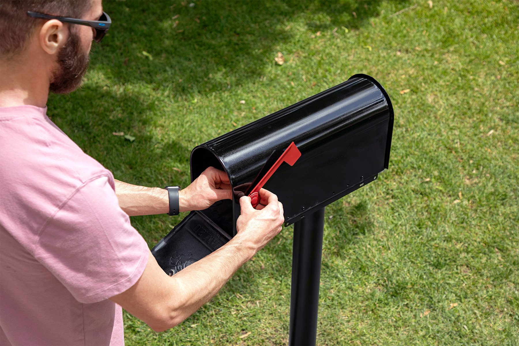 A person wearing a pink shirt and sunglasses is opening a black mailbox with a red flag. The mailbox is situated on a grassy lawn. The person is reaching inside the mailbox, possibly checking for mail.